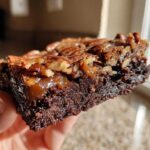 A person holding a thick, fudgy slice of pecan pie brownies showing the gooey caramel layer and pecan topping.