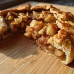 A slice cut from a rustic apple galette showing cinnamon-spiced apple filling and flaky crust, resting on a wooden cutting board.