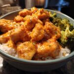 Close-up of a bowl filled with white rice, topped with saucy Bang Bang Chicken pieces and steamed broccoli florets.