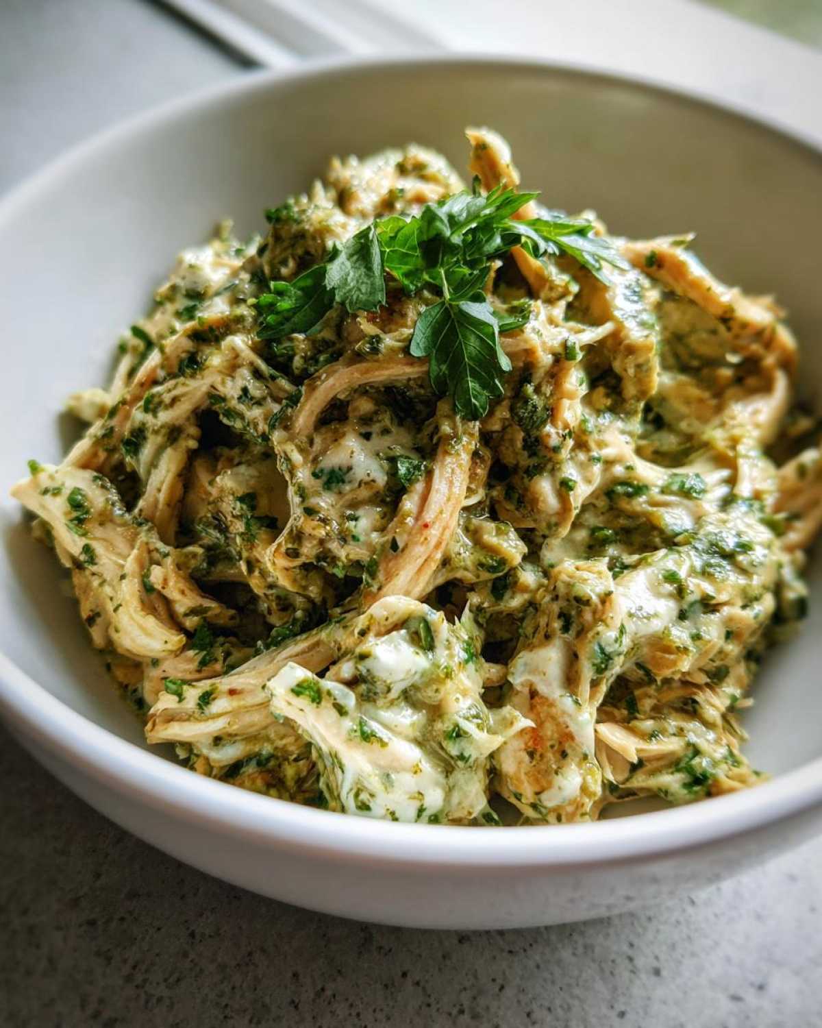 Close-up of shredded chicken coated in creamy green salsa verde sauce, garnished with parsley, ready to eat.