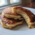 Close-up of three golden-brown Banana Pancakes stacked on a white plate, one broken open to show the fluffy interior.