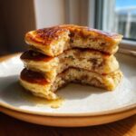 A stack of four fluffy Banana Pancakes, cut open to show the interior texture, drizzled with syrup.