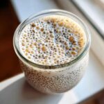 Overhead view of freshly made chia seed pudding in a glass jar, showing soaked chia seeds and a thick layer of light brown foam on top.