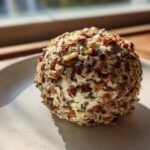 A single, round cheese ball recipe coated entirely in chopped pecans and herbs, sitting on a light plate near a window.