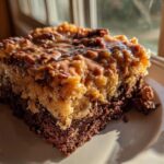 A close-up of a moist slice of german chocolate poke cake featuring a dark chocolate base and a gooey coconut-pecan topping.