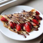Close-up of delicious Gluten-Free Chocolate Strawberry Crepes folded and filled with fresh strawberries and cream, drizzled with chocolate sauce.