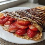A folded Gluten-Free Chocolate Strawberry Crepe filled with fresh strawberries and cream, drizzled with chocolate sauce and powdered sugar.