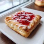 A single, flaky Gluten-Free Strawberry Cream Cheese Danishes topped with bright red strawberry filling and white icing drizzle.