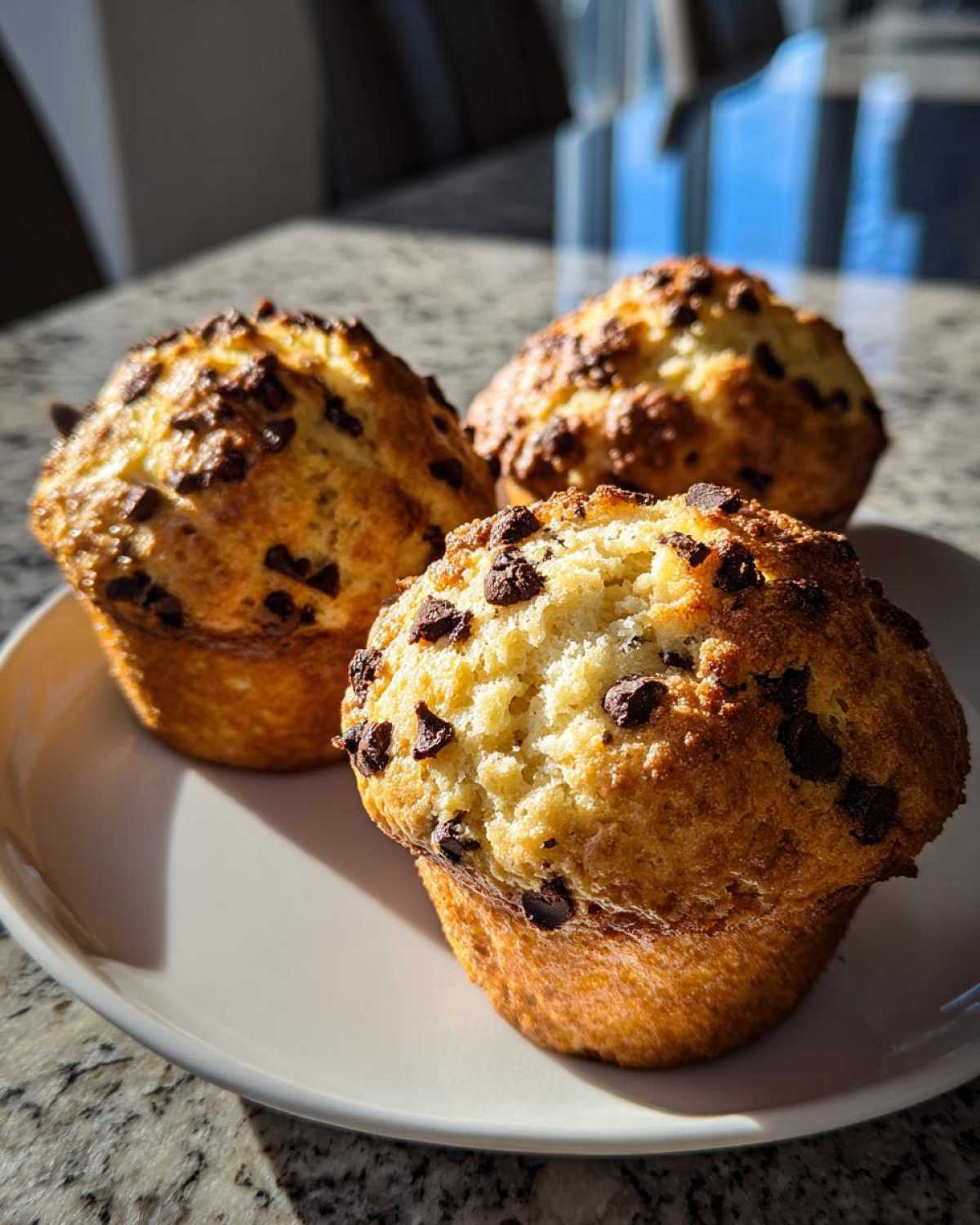 Three perfectly baked, golden brown chocolate chip muffins with visible mini chocolate chips on top, sitting on a white plate.