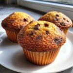 Three golden-brown chocolate chip muffins sitting on a white plate, highlighted by natural sunlight.