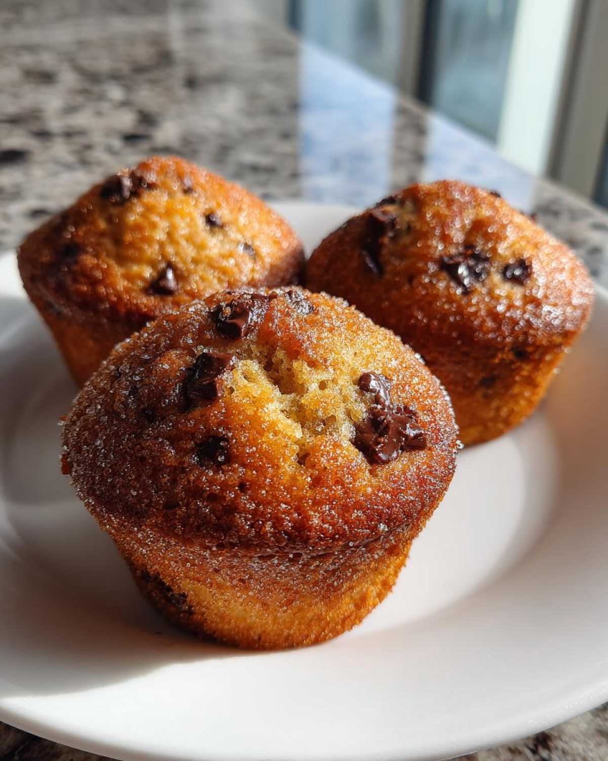 Three golden brown chocolate chip muffins dusted with sugar sitting on a white plate.