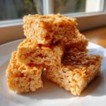 A stack of four golden, gooey crispy rice treats on a white plate, catching sunlight near a window.