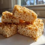 A close-up stack of golden brown, chewy crispy rice treats glistening in the sunlight on a white plate.