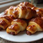 A close-up of several shiny, golden-brown pigs in a blanket appetizers stacked on a white plate.
