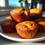 A close-up of several golden brown mini banana muffins stacked on a white plate, one has a bite taken out.
