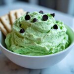 A bowl of bright green Grinch dip topped with chocolate chips, served with crackers on the side.