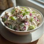A bowl filled with creamy high protein chicken salad featuring chunks of chicken, green celery, and red onion, garnished with chives.