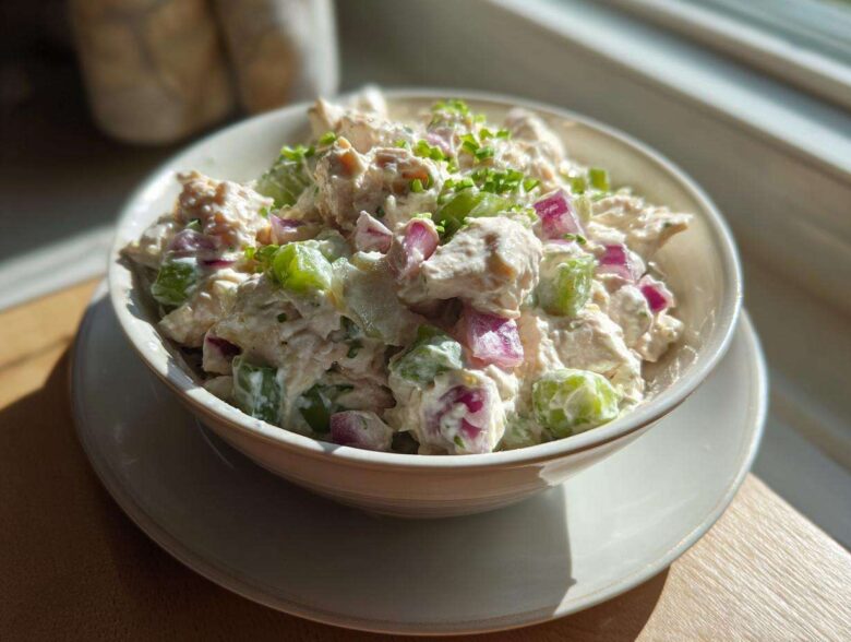 A bowl filled with creamy high protein chicken salad featuring chunks of chicken, green celery, and red onion, garnished with chives.