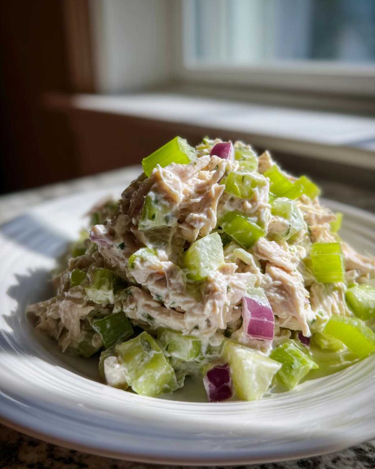 A mound of creamy high protein chicken salad mixed with large chunks of bright green celery and diced red onion, served on a white plate.