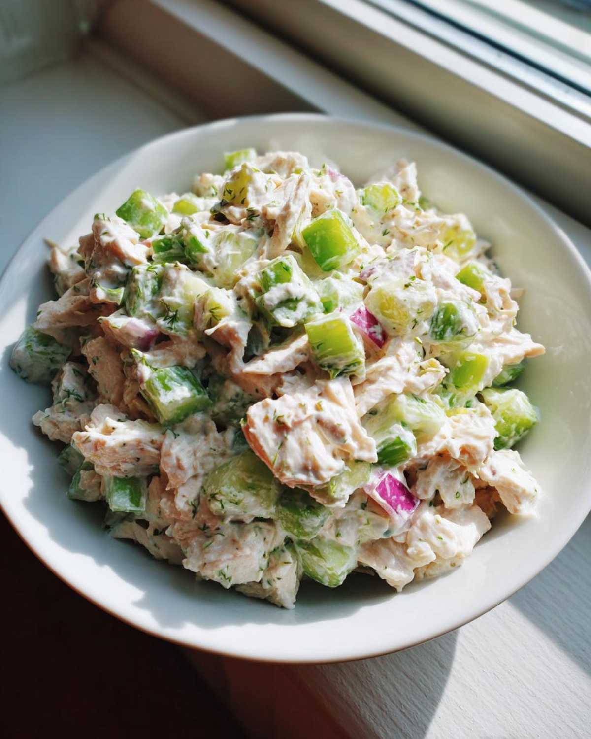 Close-up of a white bowl filled with creamy high protein chicken salad featuring chunks of chicken, green celery, and red onion.