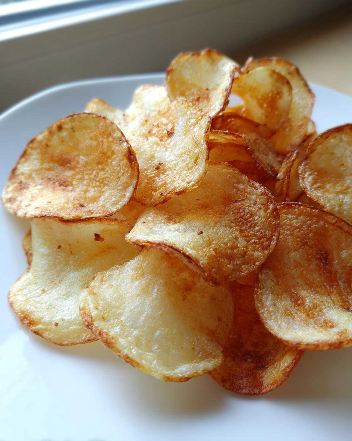 Close-up of a pile of freshly made, golden brown homemade potato chips served on a white plate.