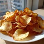 A generous pile of golden, crispy homemade potato chips served on a white plate near a window.