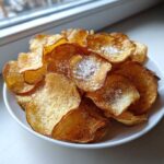 A close-up of a white bowl filled with golden brown homemade potato chips generously topped with coarse sea salt.