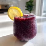 A close-up of a vibrant, deep purple Iced Blueberry Lemonade Slush garnished with a fresh lemon slice.