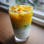 A tall glass showing the layers of an Iced Mango Coconut Matcha drink: white coconut milk base, green matcha layer, and bright yellow mango topping with ice.
