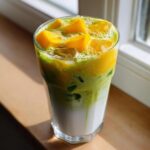 A tall glass showing layered Iced Mango Coconut Matcha with white milk, green matcha, and topped with bright yellow mango chunks.