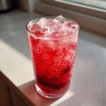 Close-up of a tall glass filled with vibrant red Iced Strawberry Hibiscus Tea Lemonade and ice cubes, showing carbonation bubbles.