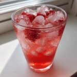 A tall, sweating glass filled with bright red Iced Strawberry Hibiscus Tea Lemonade, ice cubes, and strawberry pieces.