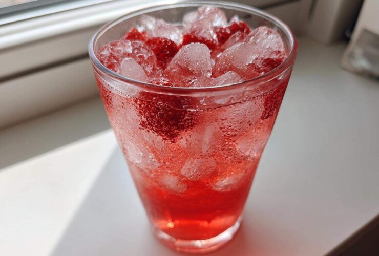 A tall, sweating glass filled with bright red Iced Strawberry Hibiscus Tea Lemonade, ice cubes, and strawberry pieces.