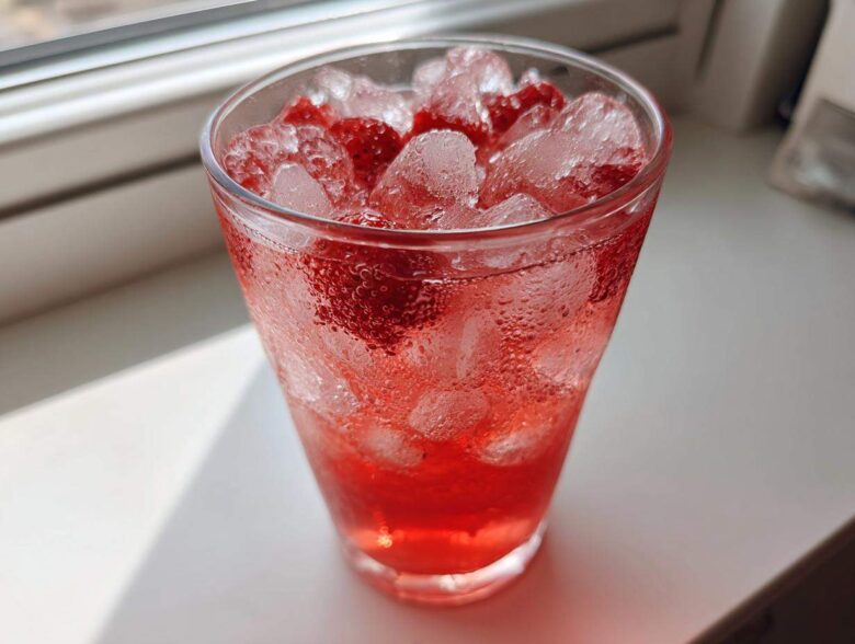 A tall, sweating glass filled with bright red Iced Strawberry Hibiscus Tea Lemonade, ice cubes, and strawberry pieces.