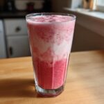 A tall glass filled with a layered Iced Vanilla Strawberry Protein Shake, showing pink and white foam.