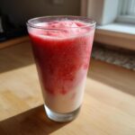 A tall glass containing a layered Iced Vanilla Strawberry Protein Shake with a pink, icy top layer and a creamy white bottom layer.