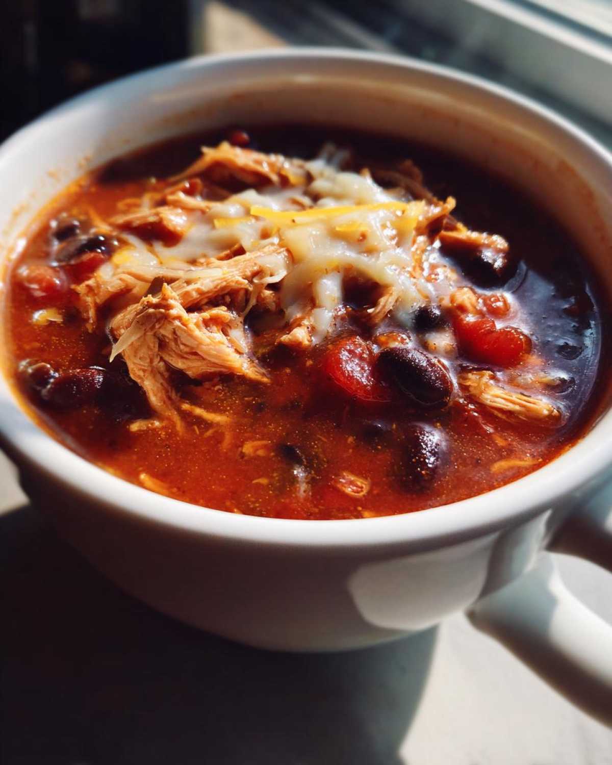 Close-up of a white bowl filled with Instant Pot Creamy Chicken Enchilada Soup, topped with shredded chicken and melted cheese.