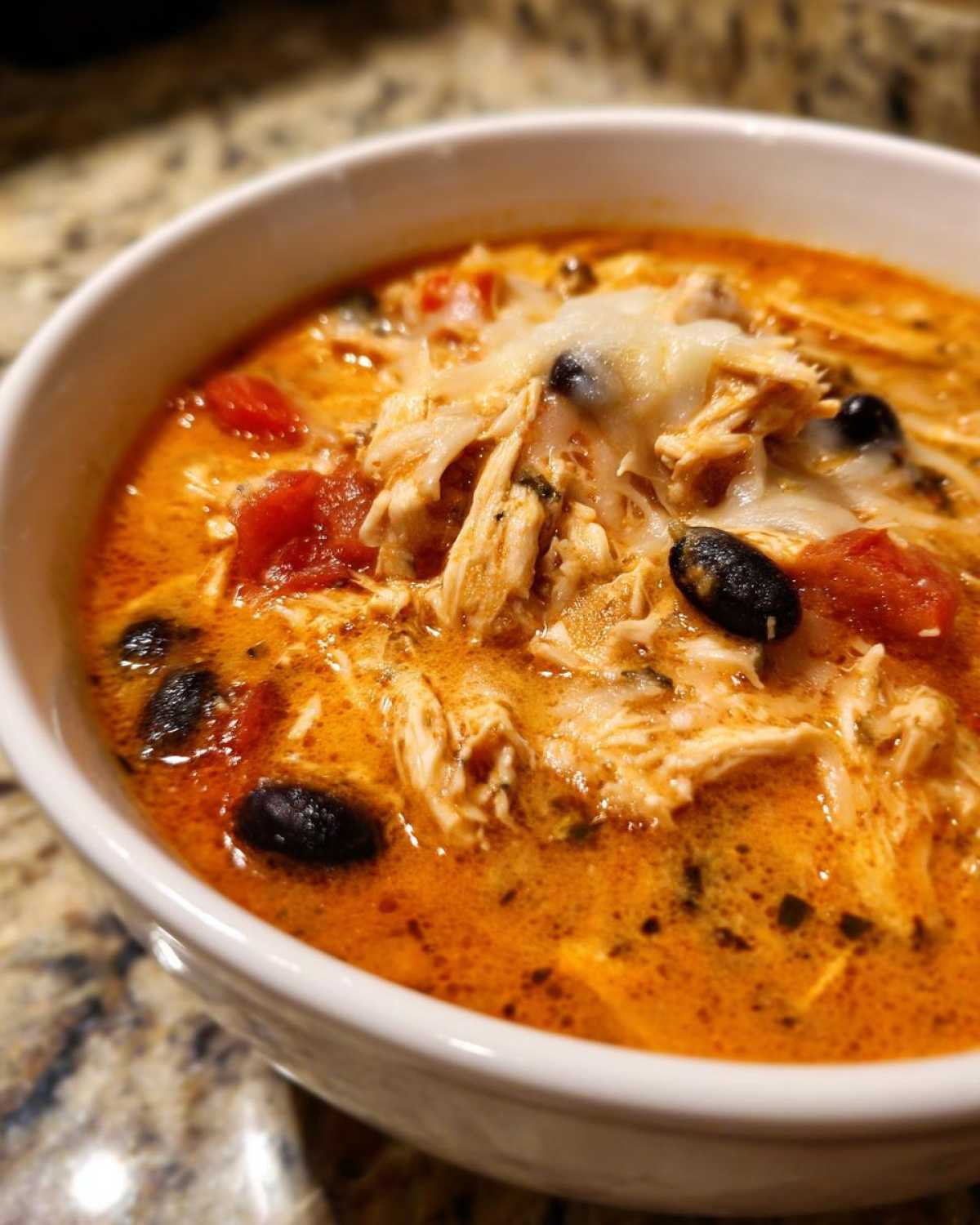 Close-up of a white bowl filled with rich, orange Instant Pot Creamy Chicken Enchilada Soup topped with shredded chicken, melted cheese, and black beans.
