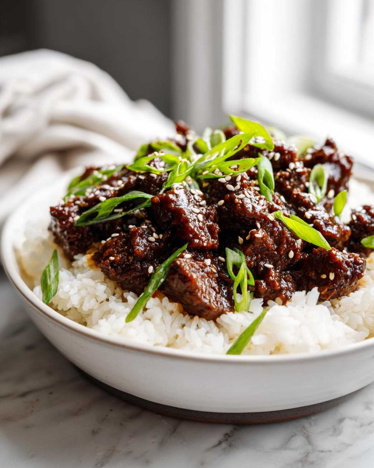 Close-up of a white bowl filled with white rice topped with saucy Instant Pot Korean Beef and garnished with green onions and sesame seeds.