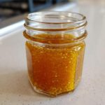 A small glass jar filled with vibrant orange Copycat Apple Cider Vinaigrette dressing, showing visible spices and seeds.