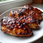 Four pieces of juicy oven baked bbq chicken covered in a thick, glossy, caramelized sauce on a white plate.