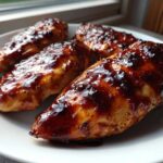 Four pieces of juicy oven baked bbq chicken breasts covered in a thick, glossy sauce served on a white plate.