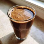 A close-up of a thick, dark brown Keto Creamy Chocolate Peanut Butter Shake in a faceted glass, sitting in sunlight.