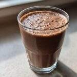 A tall glass filled with a thick, frothy Keto Creamy Chocolate Peanut Butter Shake, sitting on a light surface near a window.