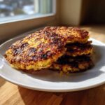 A stack of four golden-brown, crispy Keto Crispy Cauliflower Hash Browns on a white plate.