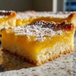 A close-up, appetizing shot of a square of bright yellow Lemon Bars topped with a dusting of white powdered sugar.