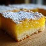 A close-up, mouth-watering shot of a single square of bright yellow Lemon Bars topped with a dusting of white powdered sugar.