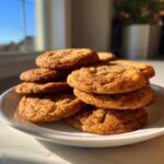 A close-up stack of golden brown maple brown sugar cookies, coated in sugar, sitting on a white plate in natural sunlight.