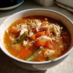 Close-up of a bowl of rich, orange-red Maryland crab soup filled with shredded crab meat, diced carrots, and green beans.