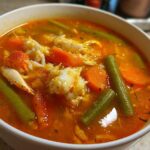 Close-up of a white bowl filled with rich, orange broth of Maryland crab soup, featuring large chunks of white crab meat, sliced carrots, and green beans.